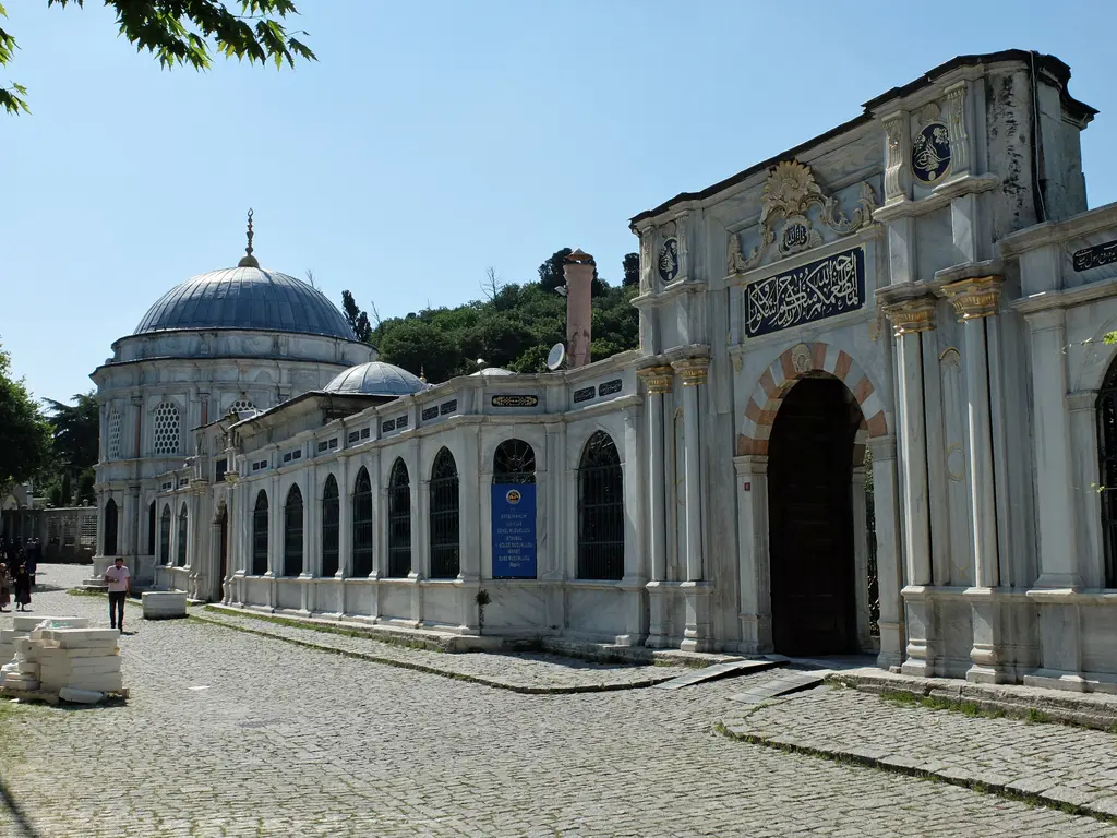 Mihrişah Valide Sultan Külliyesi'nin cephesi; kemerli taç kapı, yazılı pano, taş döşeli sokak, arkada türbe kubbesi ve bacalar.