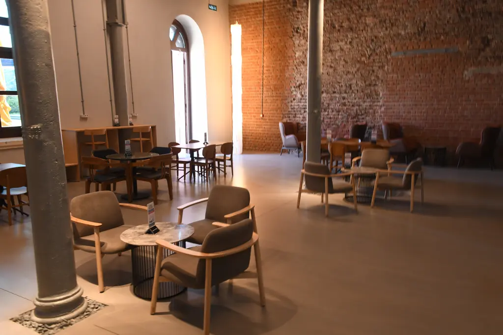 Feshane'de kafe; açık renk zeminde koltuklar ve masalar, gri demir sütun, kemerli pencere ve tuğla duvar.