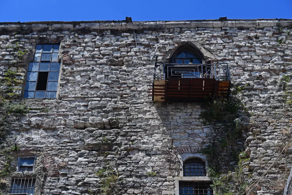 Büyük Valide Han'ın dış duvarı; kaba moloz taş ile tuğlanın almaşık örgüsü, mavi çerçeveli pencereler, kemer içine kurulmuş ahşap balkon ve duvardaki otlar.