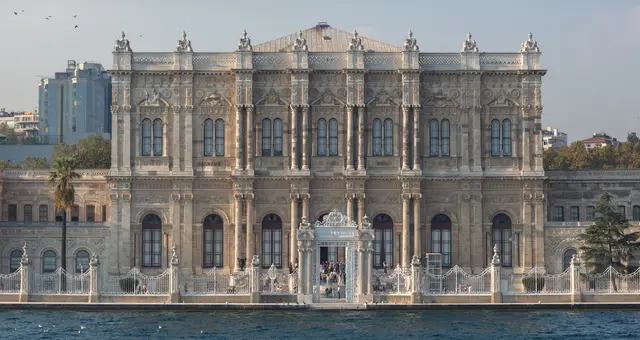 Beşiktaş'ta Dolmabahçe Sarayı; Boğaz kıyısında uzanan beyaz mermer cephe, sütunlu ve oymalı orta bölüm, önde deniz ve arkada yüksek binalar.