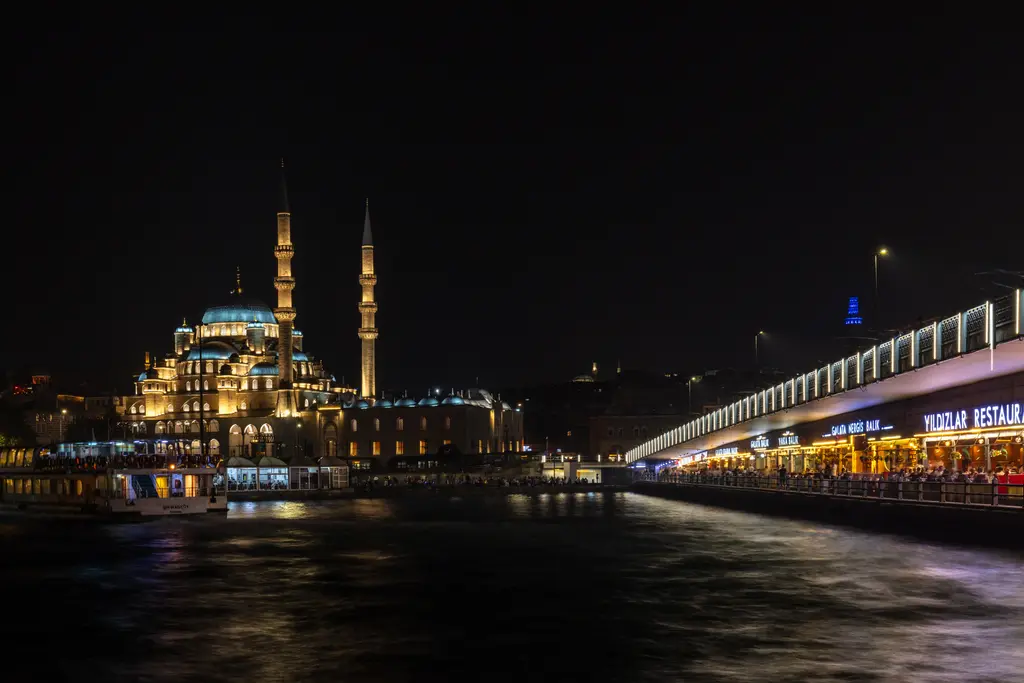 Gece Galata Köprüsü ve Yeni Cami; ışıklı köprü gövdesi, aydınlatılmış kubbe ve minareler, suya yansıyan ışıklar.