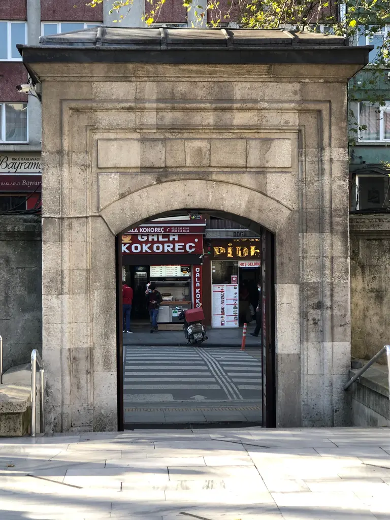 Şişli Camii'nin avlu kapısı; basık kemerli taş kapı, kapının ardında cadde ve dükkân tabelaları, önde taş döşeme.