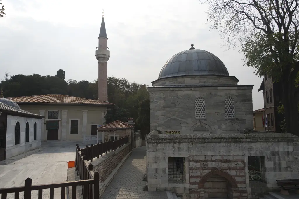 Külliyenin avlusu; ortada kubbeli kâgir türbe, arkasında minare, önde demir korkuluklu hazire ve ıslak taş döşeme.