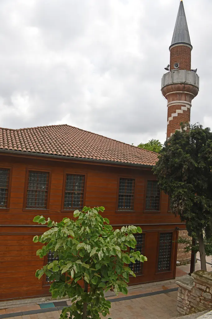 Şah Sultan Camii'nde iki katlı ahşap bina; kiremit çatı, sıralı pencereler, önde ağaç ve arkada tuğla minare.