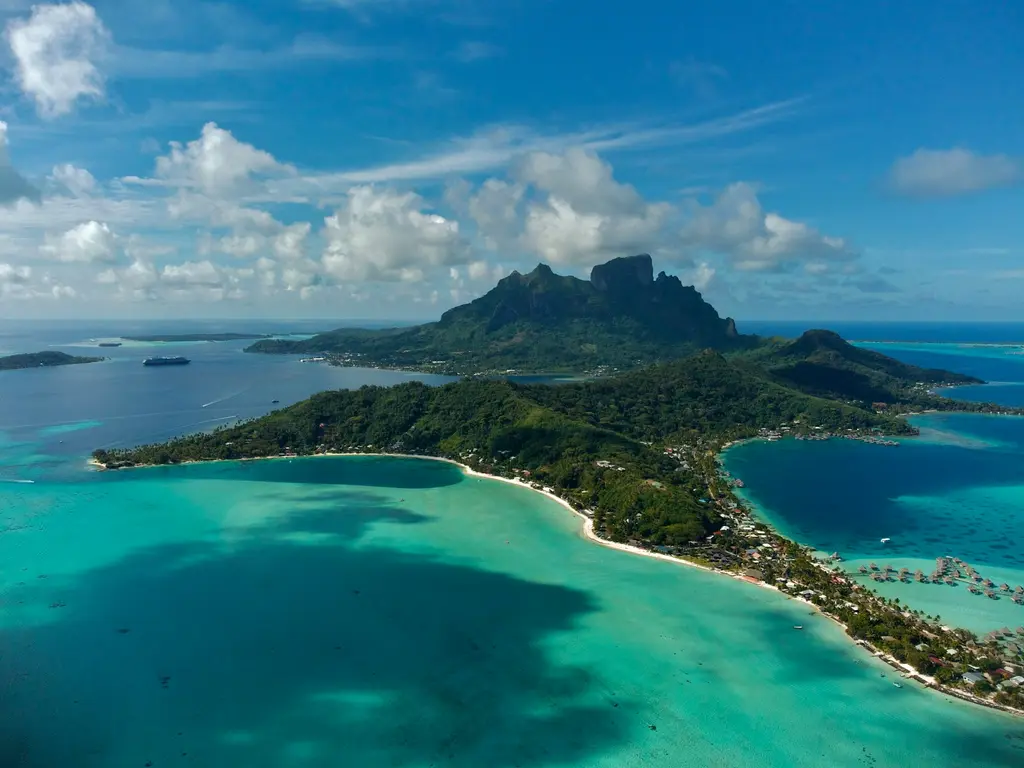 Bora Bora ve lagünündeki motuların havadan görünümü; ortada yükselen volkanik dağ, beyaz kumsallı yeşil burunlar, su üstü bungalovlar ve açıkta demirli yolcu gemisi.