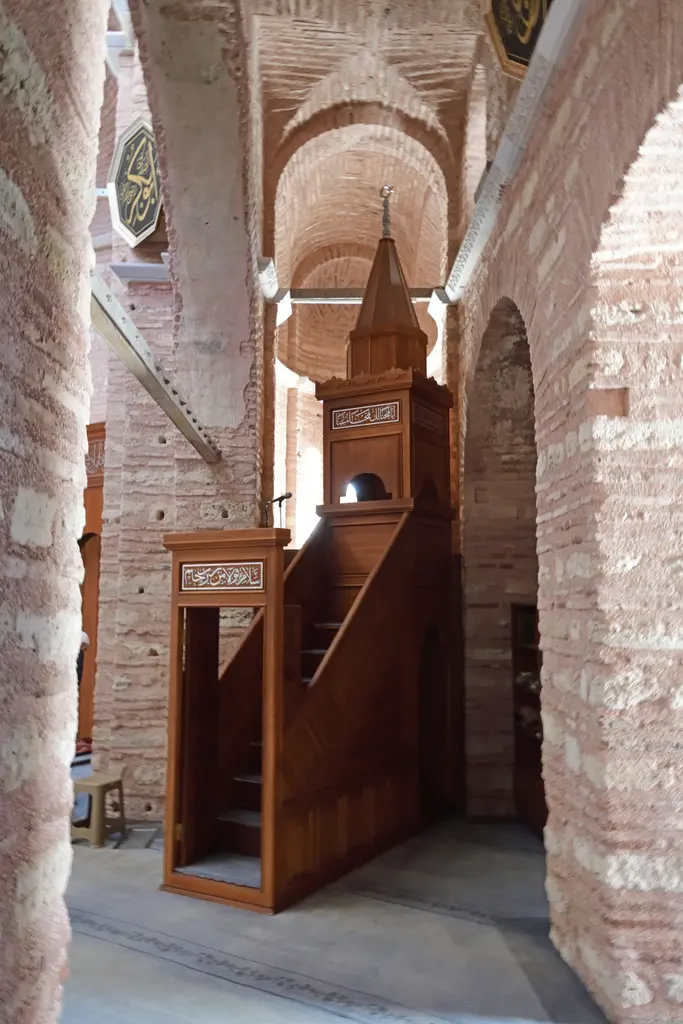 Fenari İsa Camii'nin açık renkli ahşap minberi; merdiveni, korkuluğu ve konik külahı, arkasında tuğla payeler ile kemerler, solda yeşil zeminli hat levhası.