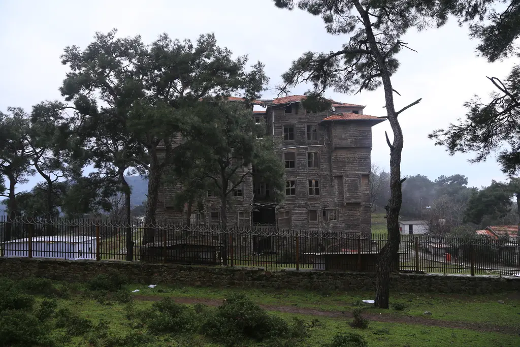 Büyükada Rum Yetimhanesi; çam ağaçlarının ardında beliren uzun ahşap bina ve önde çit ile yeşillik.