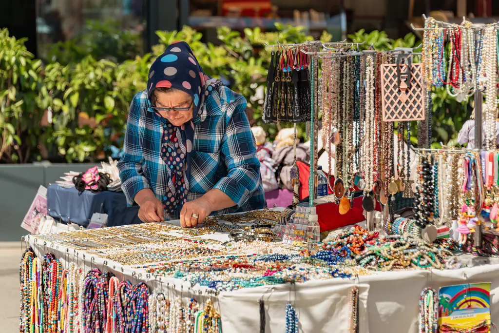 Ortaköy'de bir takı tezgâhı; renkli boncuk kolyeler, bileklikler, tezgâh başında çalışan bir kadın ve yeşil yapraklar.