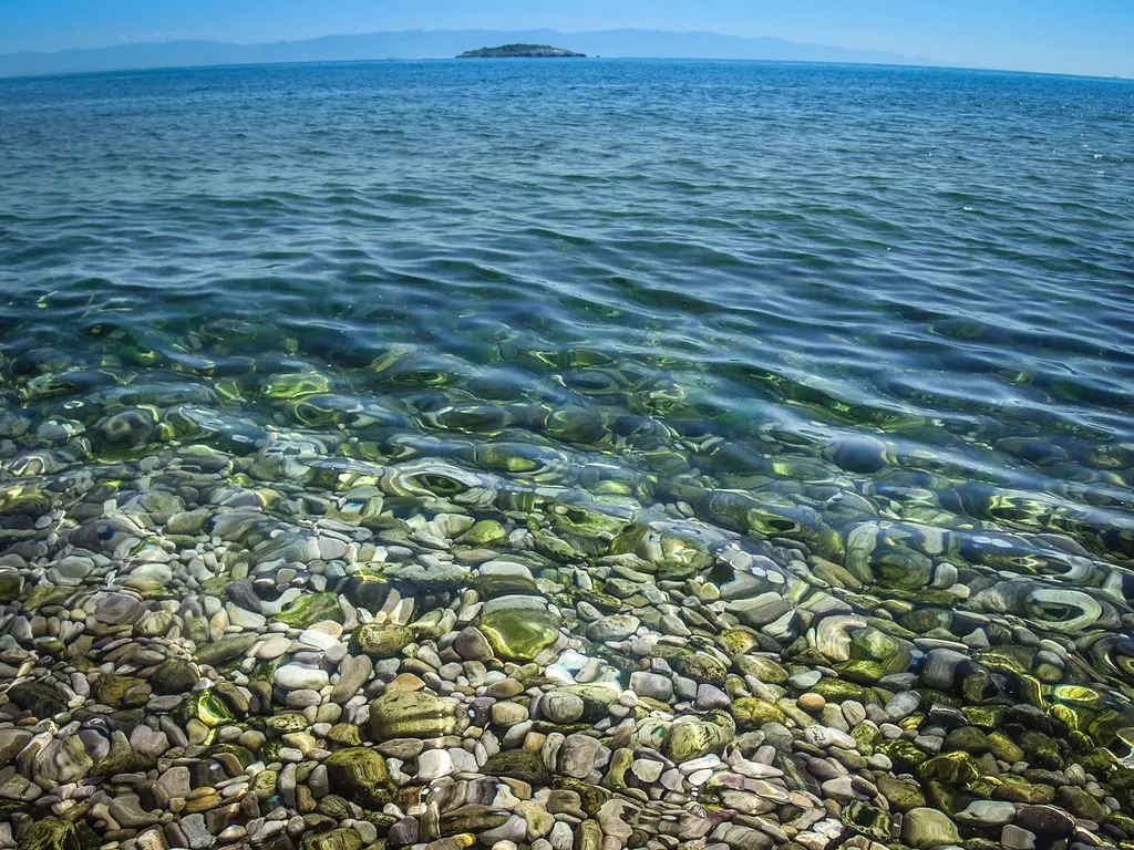 Adalar'da Tavşan Adası; berrak sığ su, dipteki çakıllar, ufukta küçük ada ve mavi gökyüzü.
