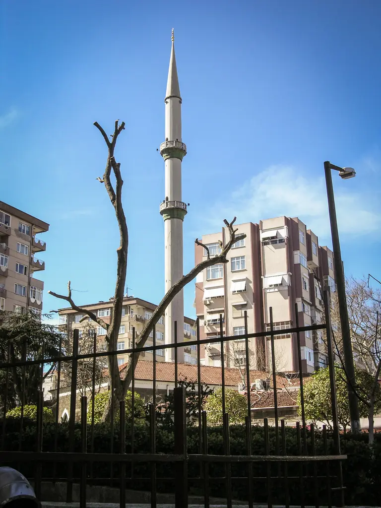 Göztepe'de Tütüncü Mehmet Efendi Camii; demir parmaklığın ardında ince beyaz minare, budanmış ağaçlar ve apartmanlar.