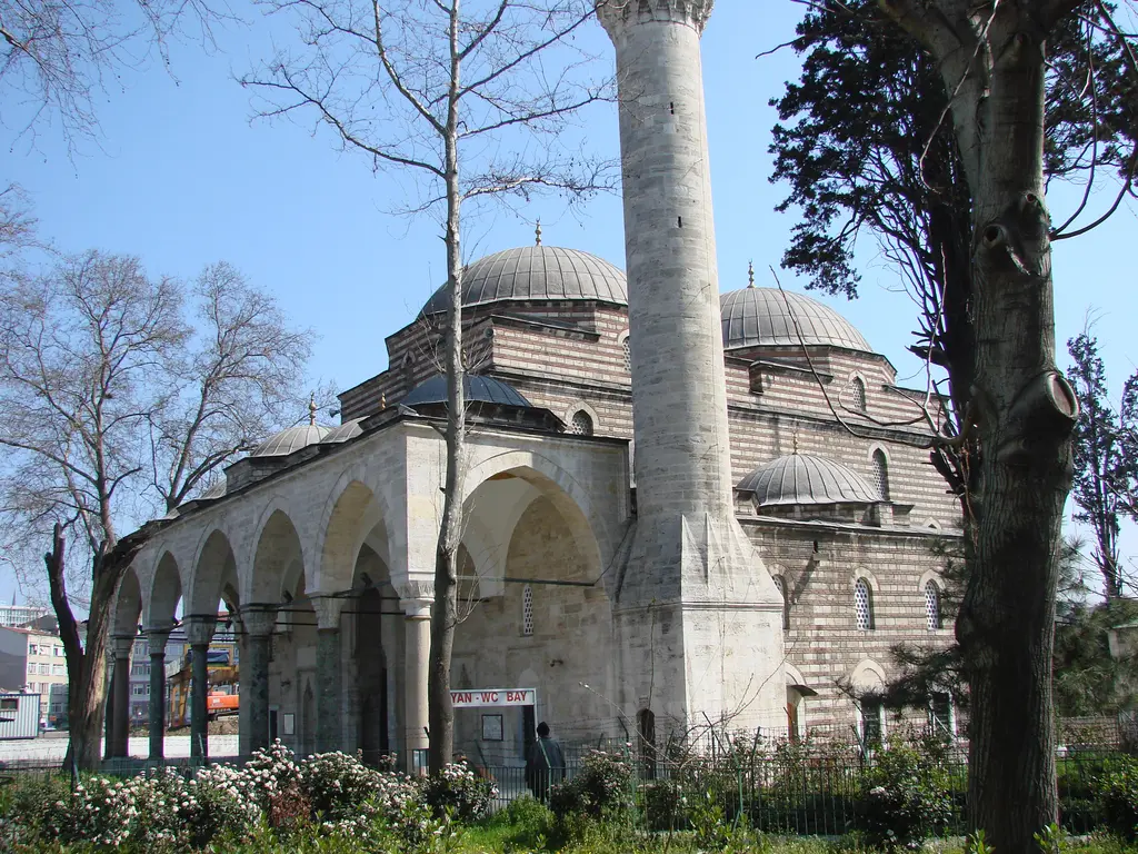 Murat Paşa Camii'ne avludan bakış; önde minarenin kalın gövdesi, arkada kemerli revak, kademeli kubbeler ve yapraksız ağaçlar.