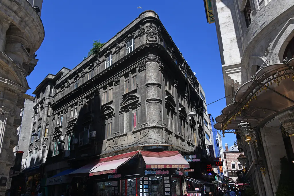 Sultanhamam Caddesi'nde köşedeki eski bina; koyu taş cephe, bezemeli pencereler, alt katta dükkânlar ve tenteler.