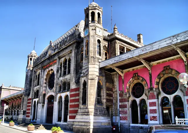 Sirkeci Garı'nın üç çeyrek açıdan görünümü; solda kubbeli saat kulesi ve taş orta blok, sağda mercan pembesi kanatta kırmızı demir kafesli gül pencereler ve ahşap saçak.