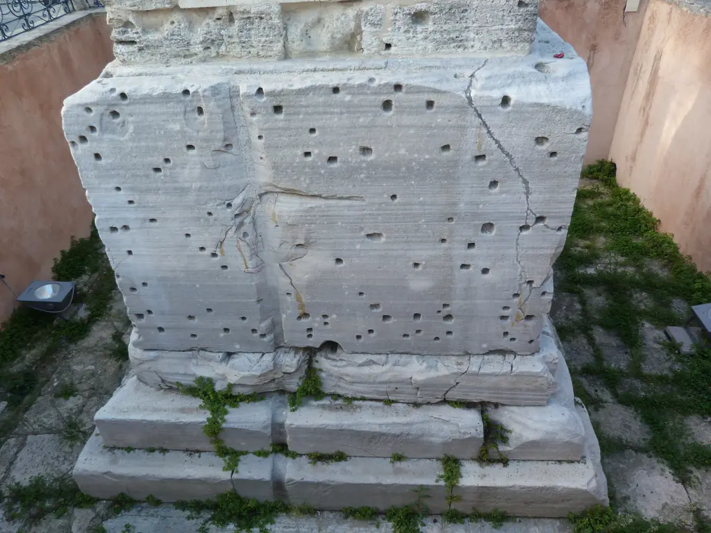 Örme Sütun'un kaidesi; kenet delikleriyle dolu mermer blok, alt basamaklar, arasında otlar ve arkada pembe sıvalı duvar.