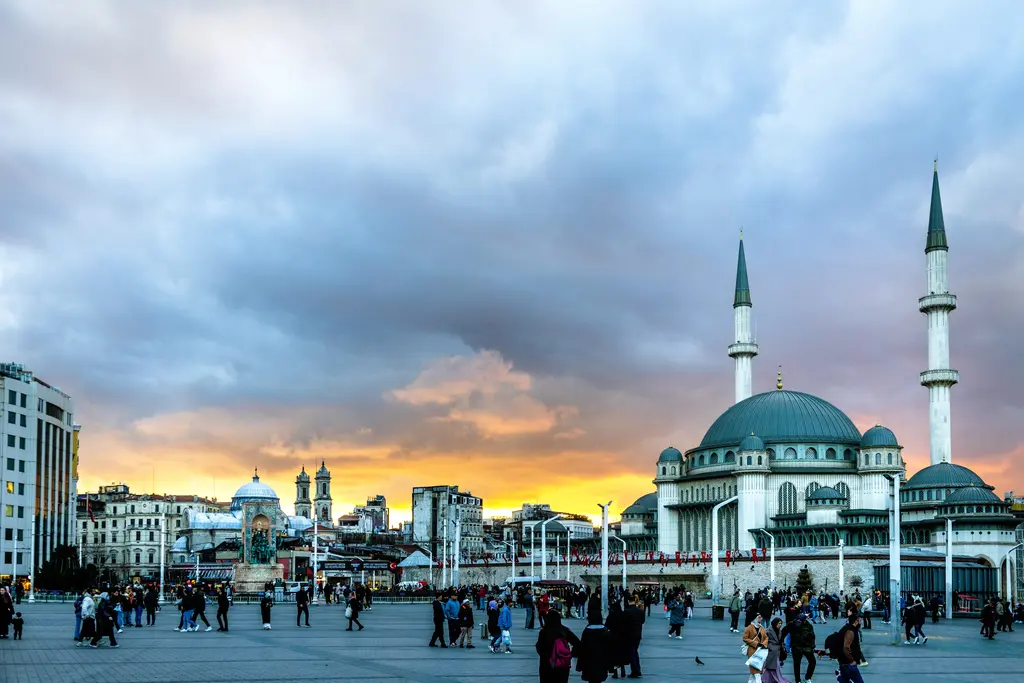 Taksim'de gün batımı; turuncu ve mavi bulutların altında Taksim Camii, meydandaki insanlar ve çevredeki apartmanlar.