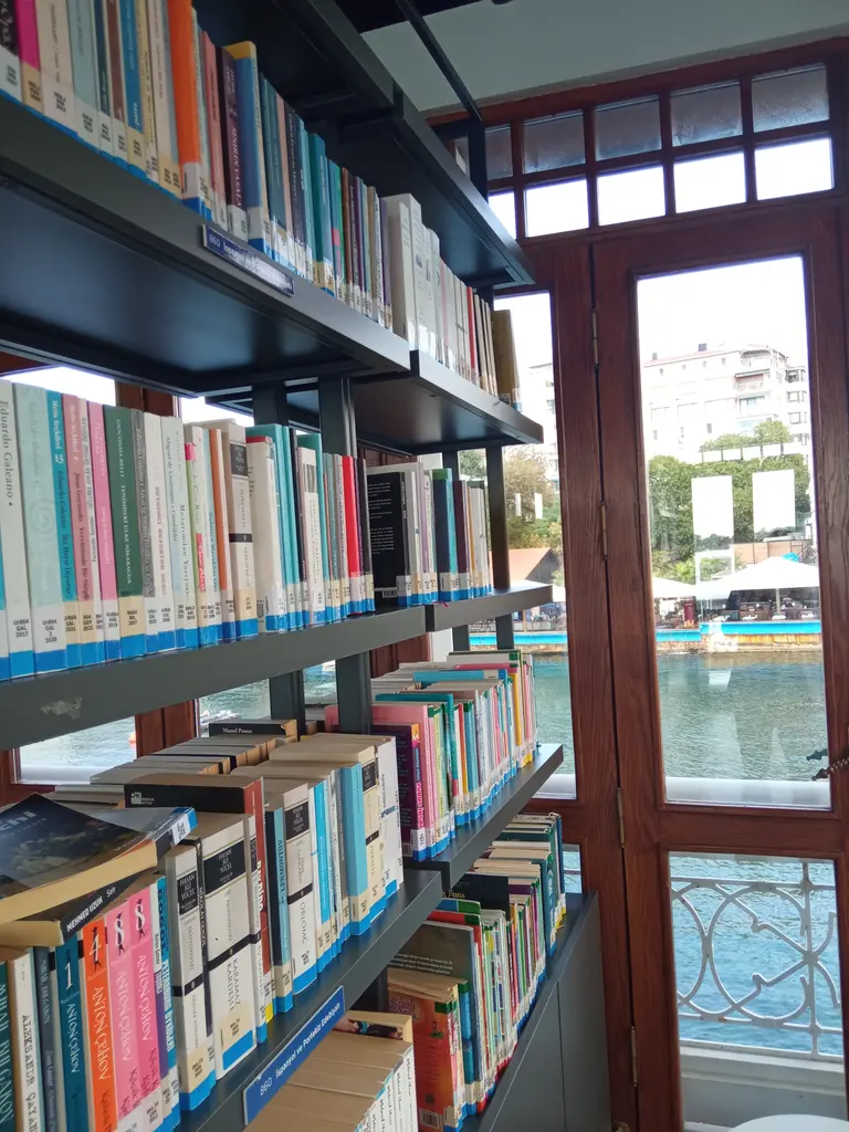 Moda İskelesi Kütüphanesi; renkli kitaplarla dolu raflar, ahşap çerçeveli pencere ve dışarıda deniz.