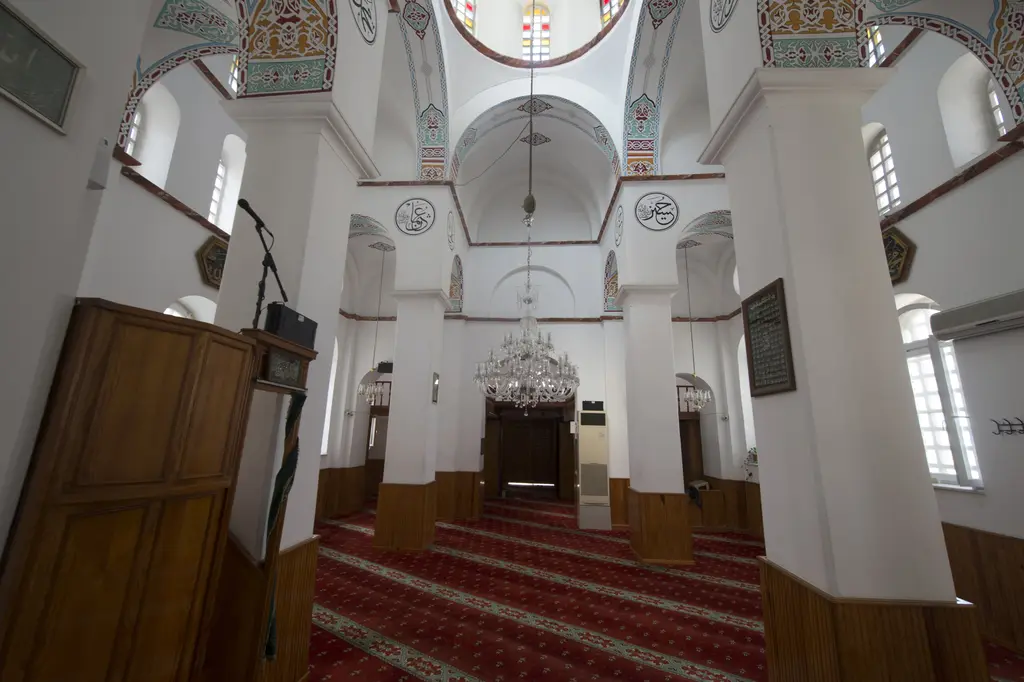 Bodrum Camii'nde giriş kapısına doğru bakış; payeler, beşik tonozlar, kubbe eteği, kristal avize, sağda hat levhası, solda ahşap kürsü ve mikrofon.