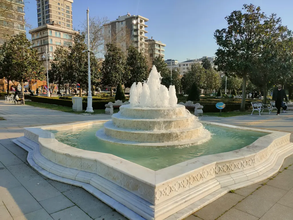 Göztepe 60. Yıl Parkı'nda mermer fıskiye; basamaklı su havuzu, taş yol, ağaçlar ve arkada yüksek binalar.