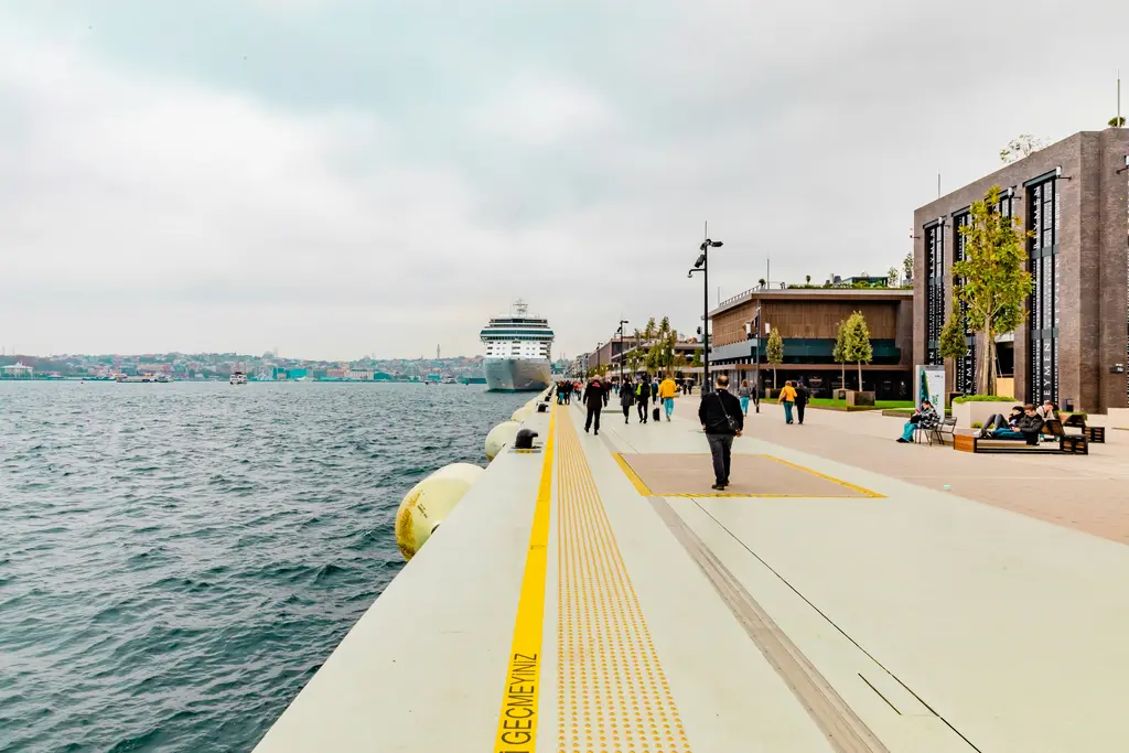 Galataport'ta Boğaz kıyısındaki yürüyüş yolu; beyaz taş rıhtım, sarı hissedilebilir yüzey şeridi, yürüyen insanlar ve uzakta bağlı yolcu gemisi.
