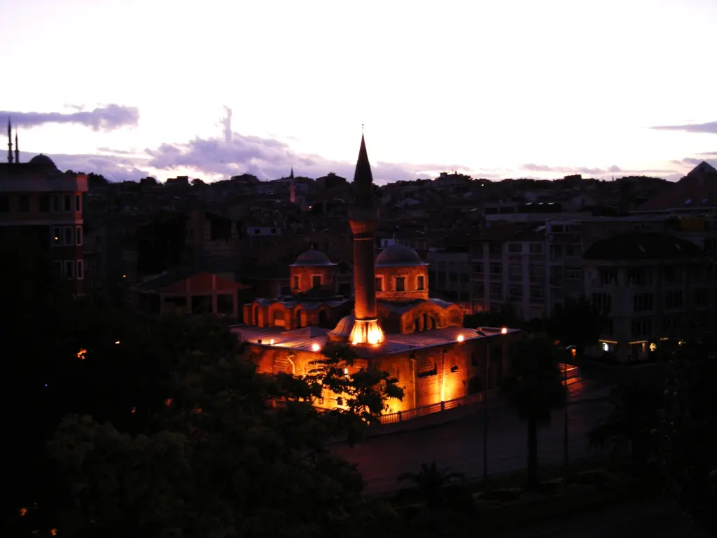 Fenari İsa Camii alacakaranlıkta; mor gri bulutlu gökyüzü altında turuncu projektörlerle aydınlatılmış gövde, boydan boya ışıklı minare ve koyu ağaç kütleleri.