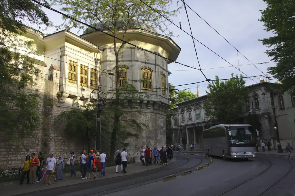İlkbaharda Alay Köşkü; yeşil ağaçlar arasında mermer salon ve sur üstündeki kanat, kaldırımda yürüyen grup ve virajı dönen bir otobüs.