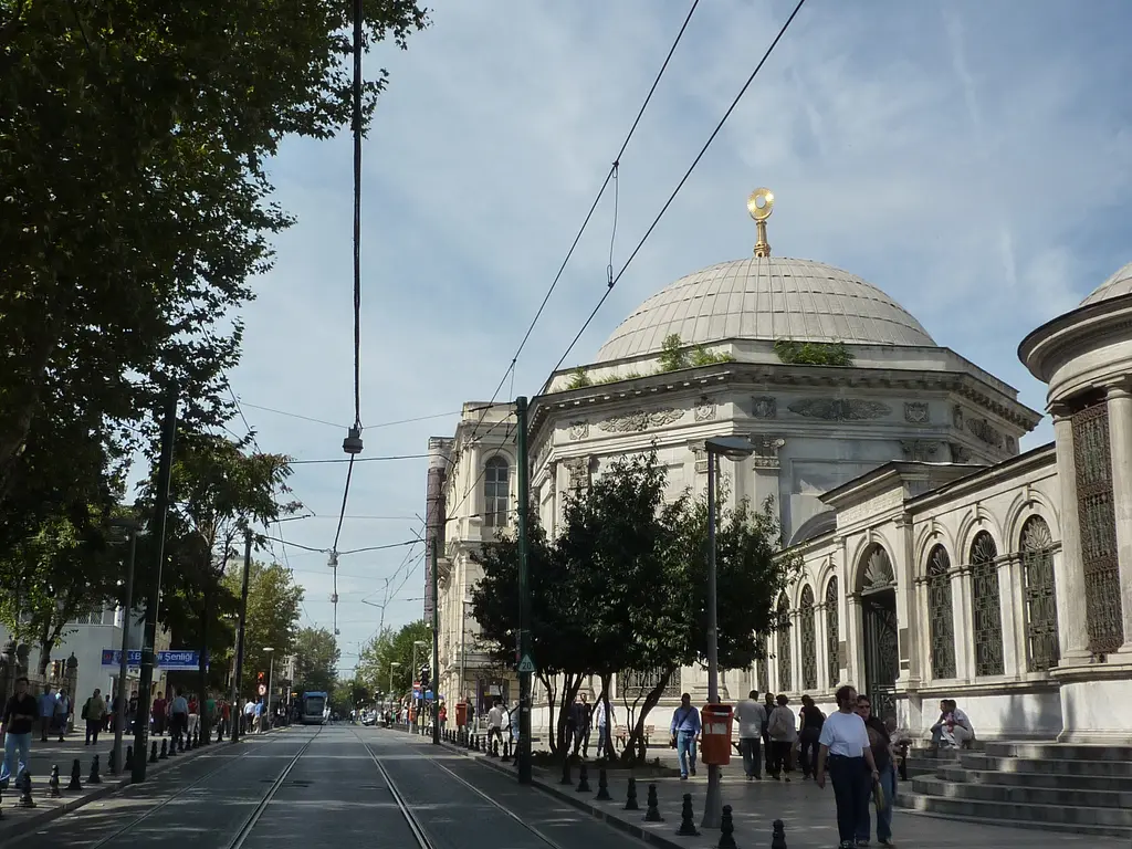 Divanyolu Caddesi'nden II. Mahmud Türbesi; tramvay rayları ve katenerler, sağda kemerli avlu duvarı, kubbeli türbe ve kaldırımda yürüyen insanlar.