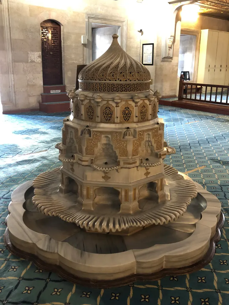 Şişli Camii'nin harimindeki mermer havuz; dilimli kenarlı tekne, oymalı ve yaldızlı kubbeli fıskiye, çevrede halı ve mahfil.