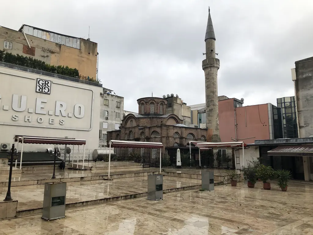 Bodrum Camii yağmurdan ıslak mermer meydandan; ortada tuğla gövde ve kubbe, sağda tam boy minare, solda dev bir ayakkabı reklamı ve çizgili tenteler.