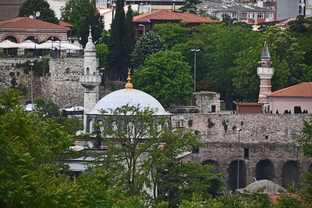 Zeyrek Sarnıcı'nın çevresi; ağaçlar arasında kurşun kubbeli küçük bir cami, minare, arkada evler ve önde sarnıcın kemerli duvarı.