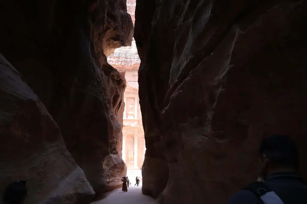 Petra'da iki yanı yüksek kaya duvarlarıyla çevrili dar kanyon geçidi; kumlu zeminde yürüyen küçük insan siluetleri ve kanyonun sonunda görünen aydınlık açıklık.