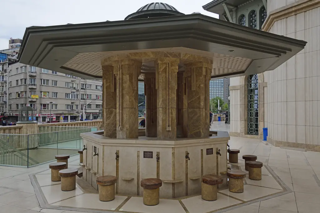 Taksim Camii'nin şadırvanı; geniş saçaklı çatı, taş sütunlar arasında hazne, çevresinde alçak oturma taşları ve arkada cami cephesi.