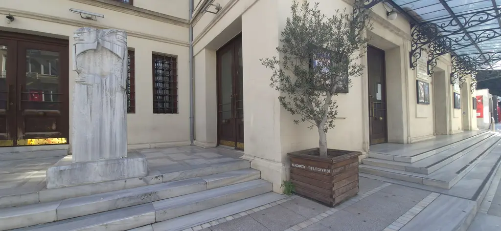 Süreyya Operası'nın girişinde mermer heykel; basamaklar, saksıda zeytin ağacı, ahşap kapılar ve demir saçak.