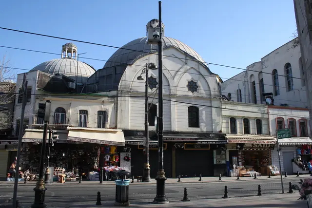 Çemberlitaş Hamamı'na sokaktan bakış; kurşun kaplı büyük ve küçük iki kubbe, kemerli alınlık, altta sıra dükkânlar ve önde sokak lambaları.