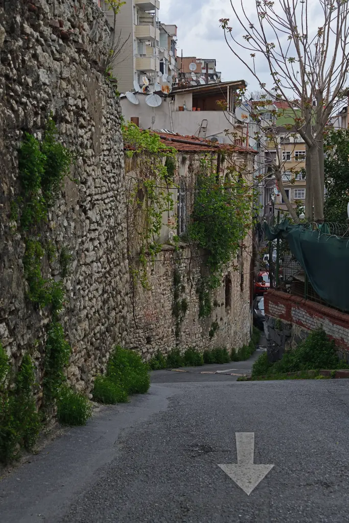 İmrahor İlyas Bey Camii'nin yanındaki dar sokak; solda sarmaşıklı iri taş duvar, sağda konutlar ile yeşil şantiye filesi, aşağı inen asfalt yol ve yol oku.