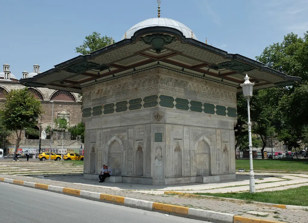 Tophane Çeşmesi; geniş saçak, kare gövde, nişler ve kabartmalar, önde taş döşeli meydan ve kaide boyunca oturan insanlar.