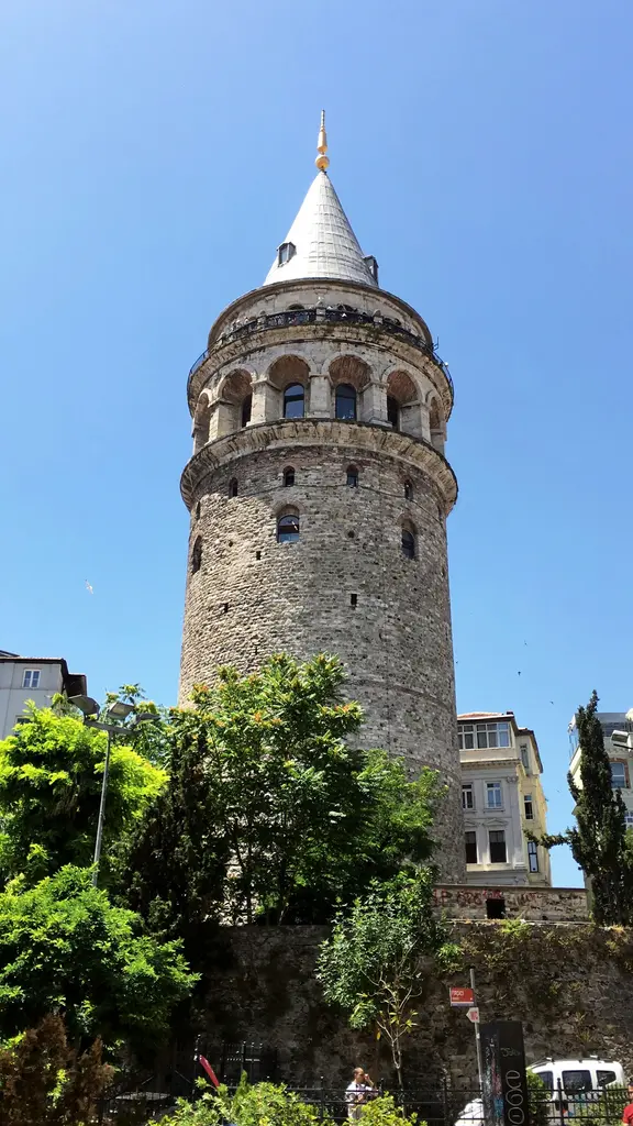 Galata Kulesi aşağıdan; mavi gökyüzüne yükselen taş gövde, kemerli pencere kuşağı, sivri külah ve önde yeşil ağaçlar.