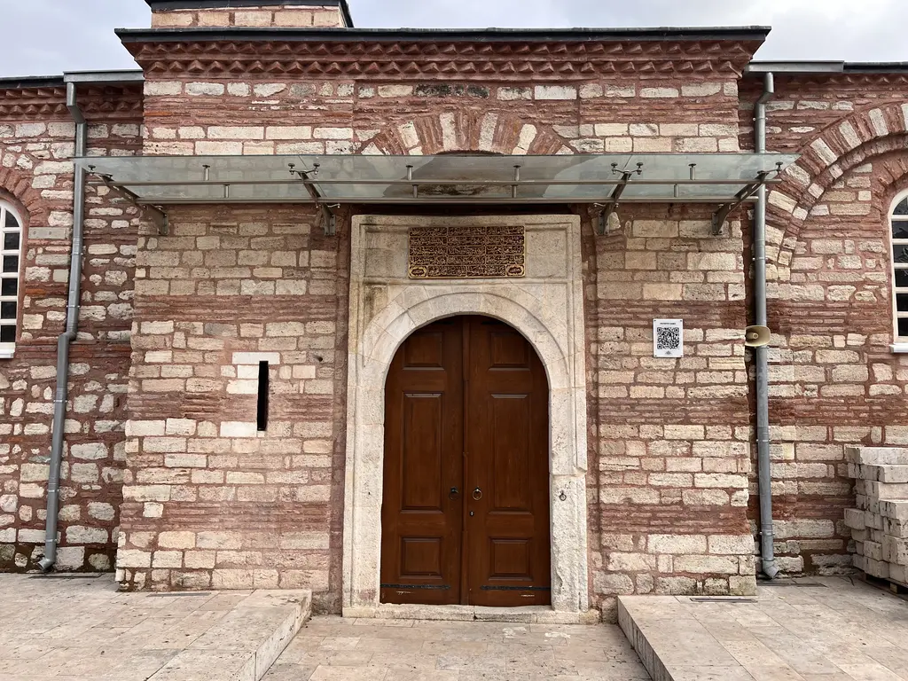 Fethiye Camii'nin giriş kapısı; tuğla taş örgülü duvarda kemerli açıklık, çift kanatlı ahşap kapı, üstünde saçak ve yanında bilgi levhası.