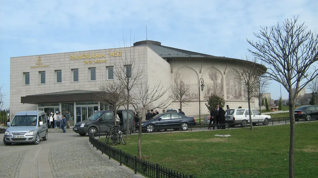 Müzenin ön cephesi ve otoparkı; uzun beyaz bina, girişte bekleyen ziyaretçiler, önünde minibüs ile otomobiller ve yapraksız ağaçlar.