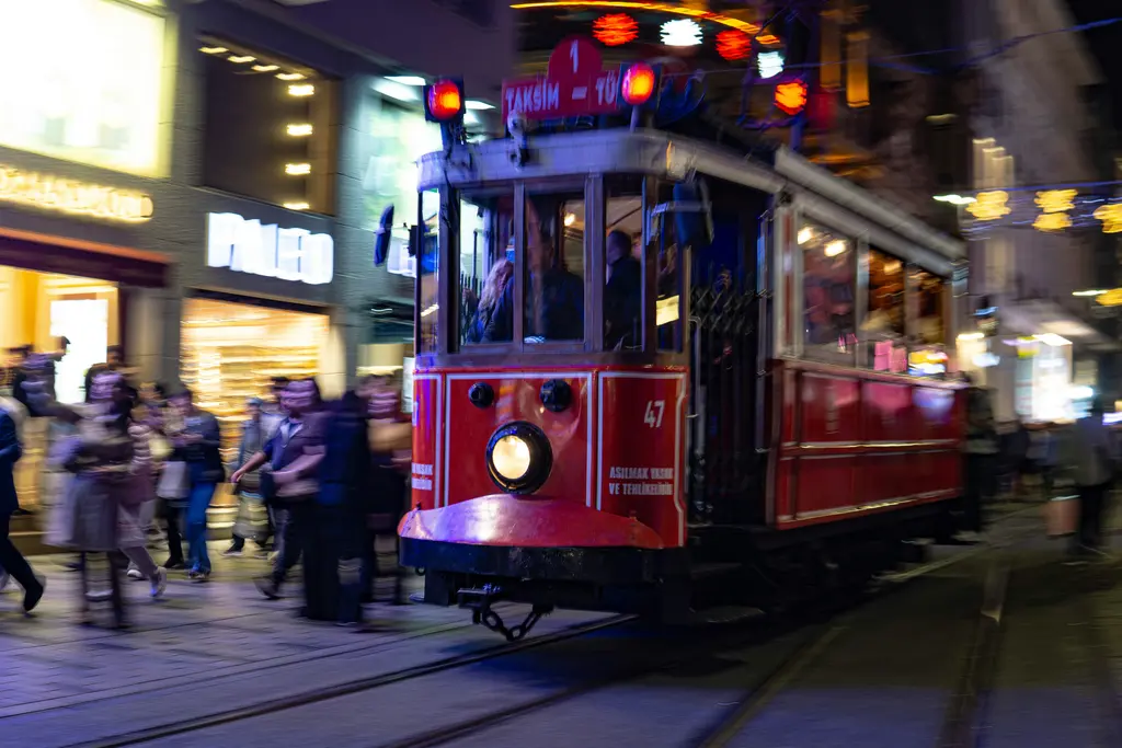 Gece İstiklal Caddesi'nde ışıkları yanan kırmızı tramvay; önünde yürüyen insanlar, iki yanda aydınlatılmış vitrinler.