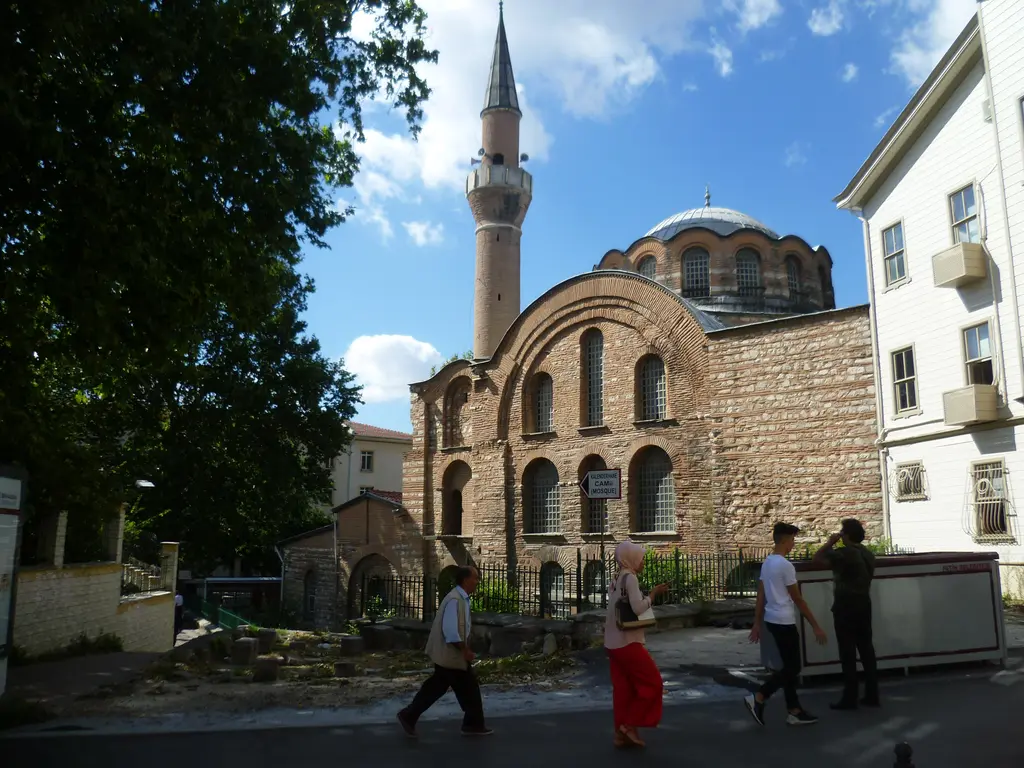 Kalenderhane Camii'ne sokaktan bakış; tuğla bantlı cephe, iki sıra kemerli pencere, arkada kubbe ve minare, önde yürüyen insanlar.