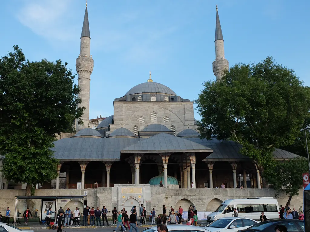 Mihrimah Sultan Camii; mavi gökyüzü altında kubbe ve iki minare, önünde yeşil ağaçlar ve meydan.