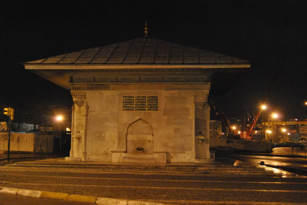 Gece III. Ahmed Çeşmesi; sarı ışıkla aydınlatılmış mermer gövde, karanlık gökyüzü ve meydandaki lambalar.