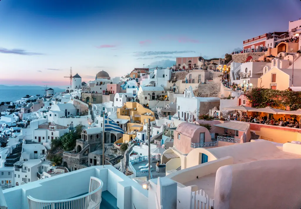 Yunanistan'da Santorini'nin Oia kasabası alacakaranlıkta; kayalığa kurulu beyaz badanalı evler, yel değirmeni, kubbeler ve kalabalık teras kafeleri.