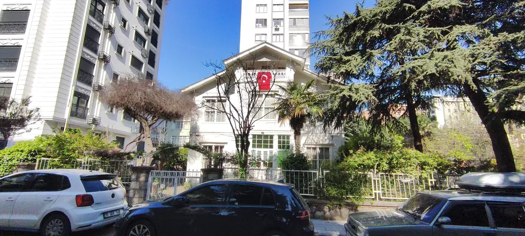 Göztepe'de Ömerpaşa Caddesi'nde konak; Türk bayraklı beyaz ahşap yapı, bahçesinde ağaçlar ve önünde park etmiş arabalar.