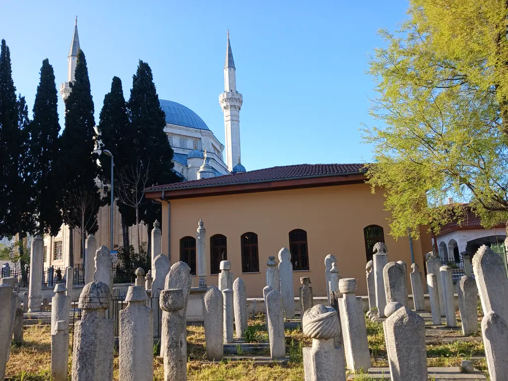 Hazirede sıra sıra duran mermer mezar taşları; selviler arasında kiremit çatılı türbe ve arkada mavi kubbeli yeni cami.
