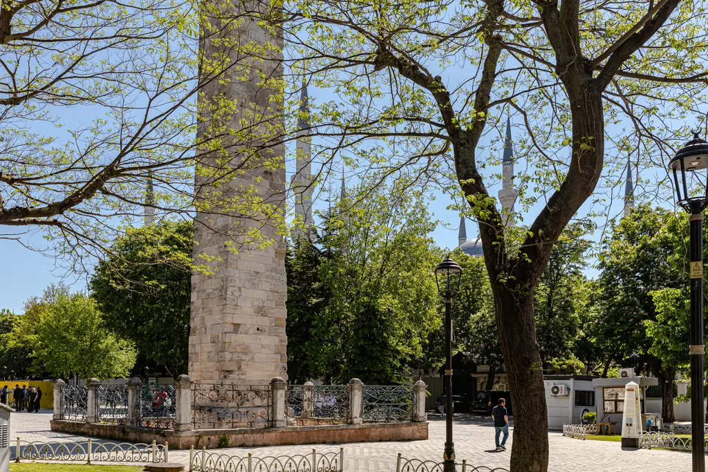 Bahar ağaçlarının arasında Örme Sütun; önde yeşil yapraklı dallar, parmaklık, sokak lambası ve meydanda yürüyen insanlar.