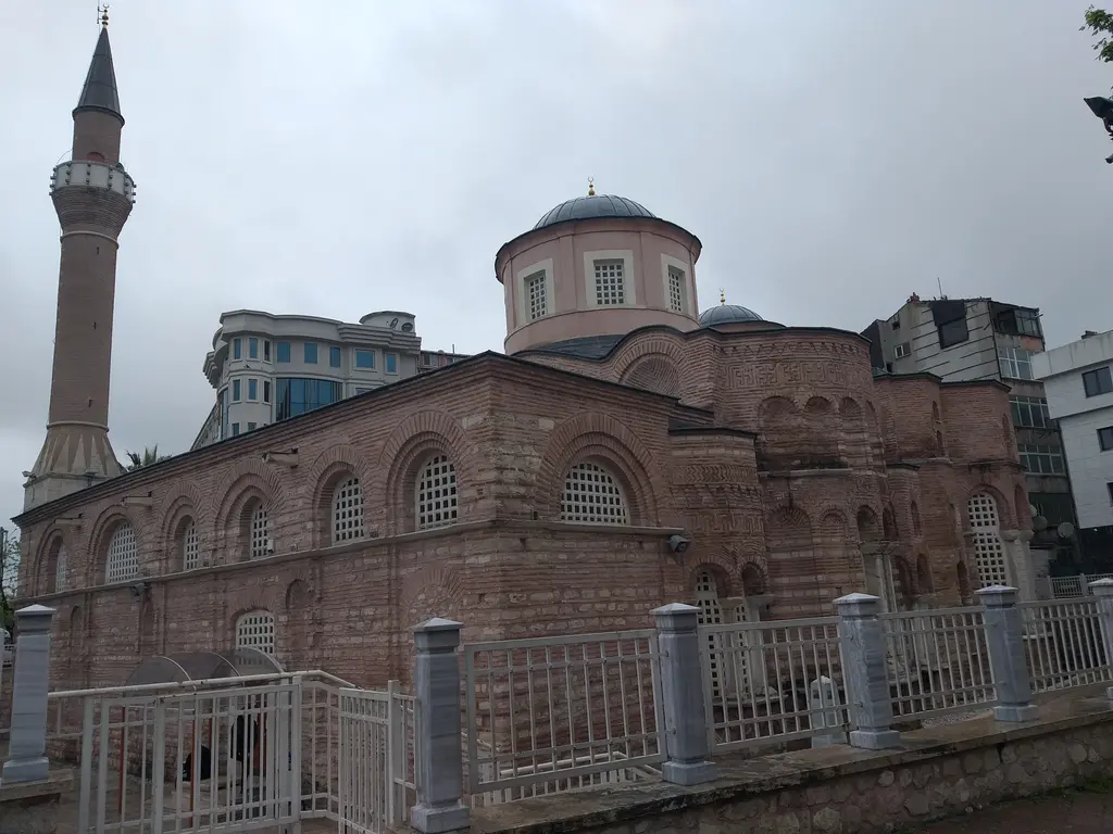 Fenari İsa Camii'ne batıdan bakış kapalı havada; solda ince minare, ortada pembe beyaz kubbe kasnağı, sağa uzanan tuğla gövde ve önde beyaz demir korkuluk.
