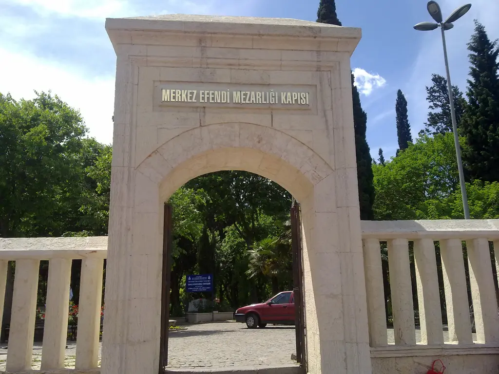 Mezarlığın kemerli taş kapısı; üstünde mezarlığın adını taşıyan levha, iki yanda alçak duvar, arkada selviler ve park eden araçlar.