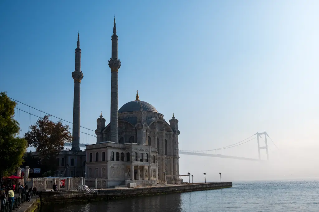 Sabah ışığında Ortaköy Camii; pusun içinden görülen köprü, kıyıdaki cami ve minareler, önde ağaçlar ve rıhtım.