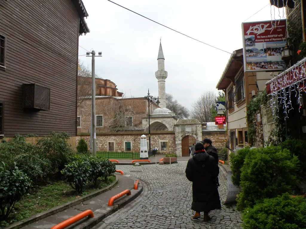 Küçük Ayasofya Mahallesi'nde kış günü; solda koyu ahşap Osmanlı evleri, sağda pansiyon ve halı dükkânları, arkada caminin taş avlu duvarı, kemerli kapısı ve minaresi.